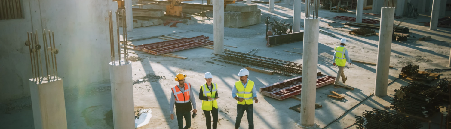 Aerial view of construction workers in hard hats and safety vests walking through a commercial building site with concrete columns.