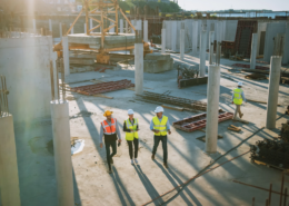 Aerial view of construction workers in hard hats and safety vests walking through a commercial building site with concrete columns.