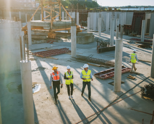 Aerial view of construction workers in hard hats and safety vests walking through a commercial building site with concrete columns.