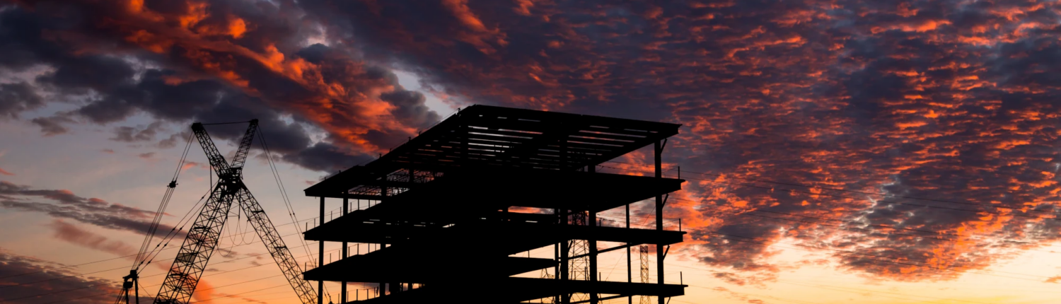 Silhouette of a commercial building under construction with a crane against a dramatic sunset sky.