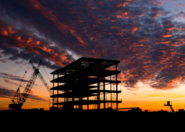 Silhouette of a commercial building under construction with a crane against a dramatic sunset sky.